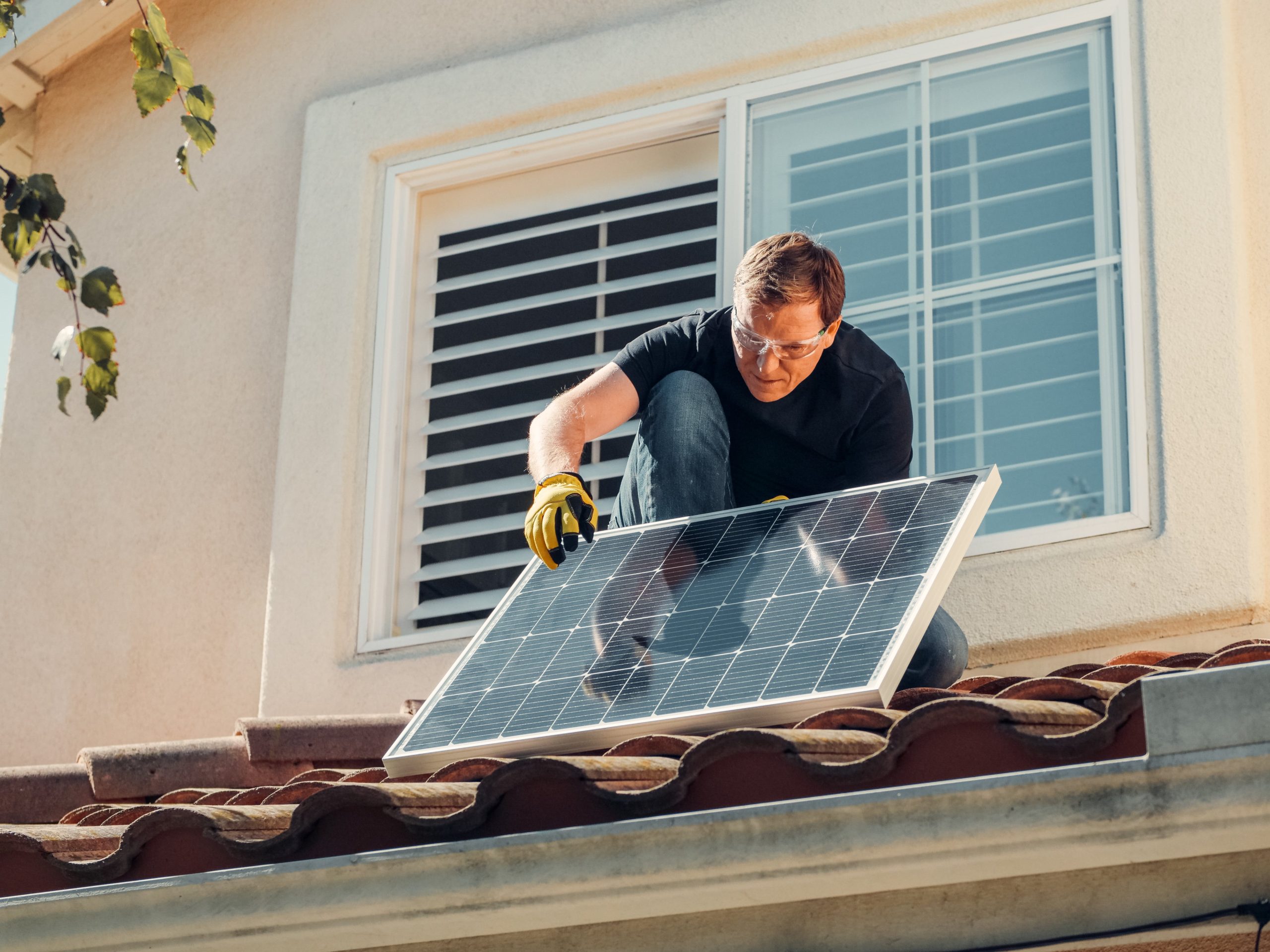 Zdjęcie przedstawia mężczyznę montującego panel fotowoltaiczny na dachu domu jednorodzinnego.